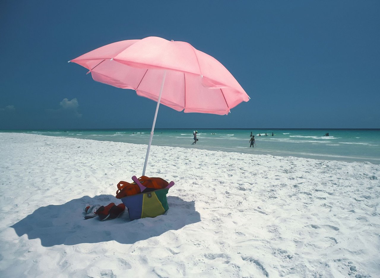 Pink beach umbrella shading a colorful bag on white sand beside turquoise water.