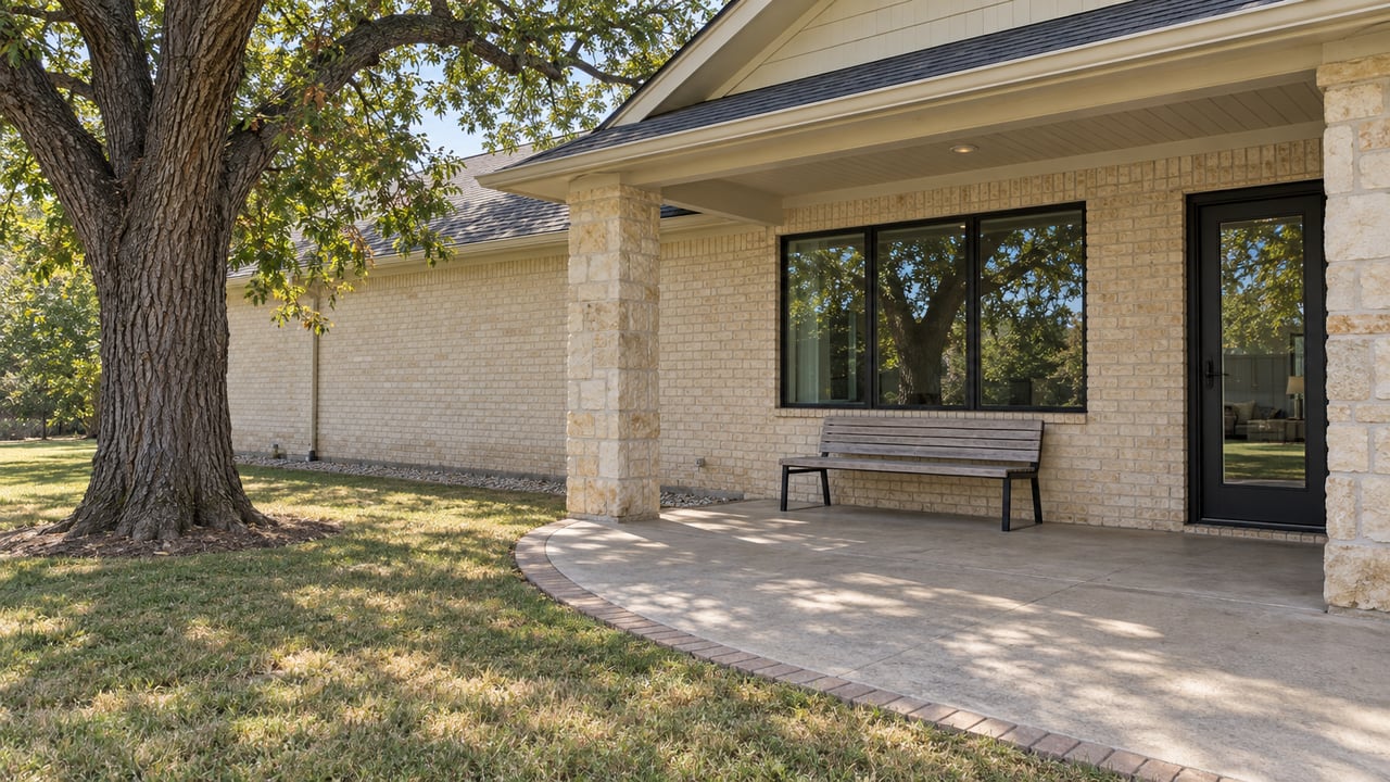 A mature pecan tree beside a dry backyard lawn and covered brick-and-limestone patio in clear morning light.