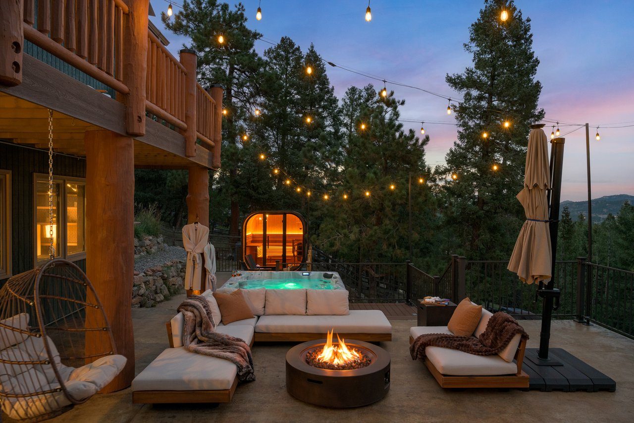 Wooden deck with a hot tub, glowing sauna, and fire pit beneath string lights.