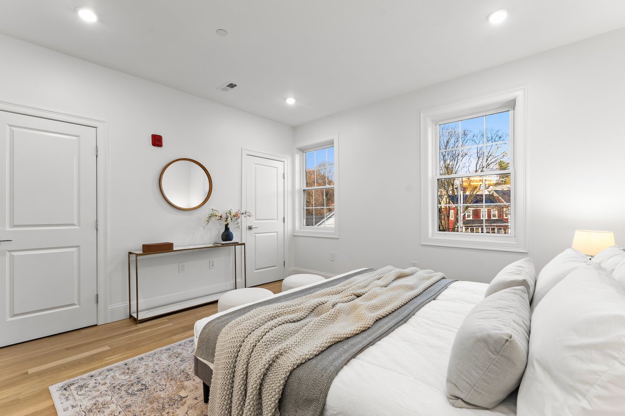 Staged bedroom with a white bed, layered blankets, hardwood floors, and two windows overlooking a neighboring house.