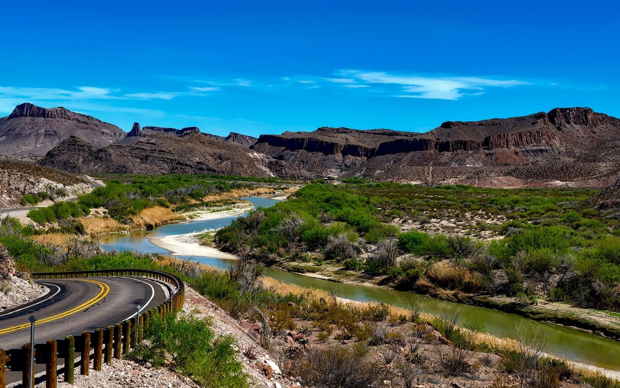  A high-angle view of a winding road with a guardrail on the left, curving through a rugged desert landscape.