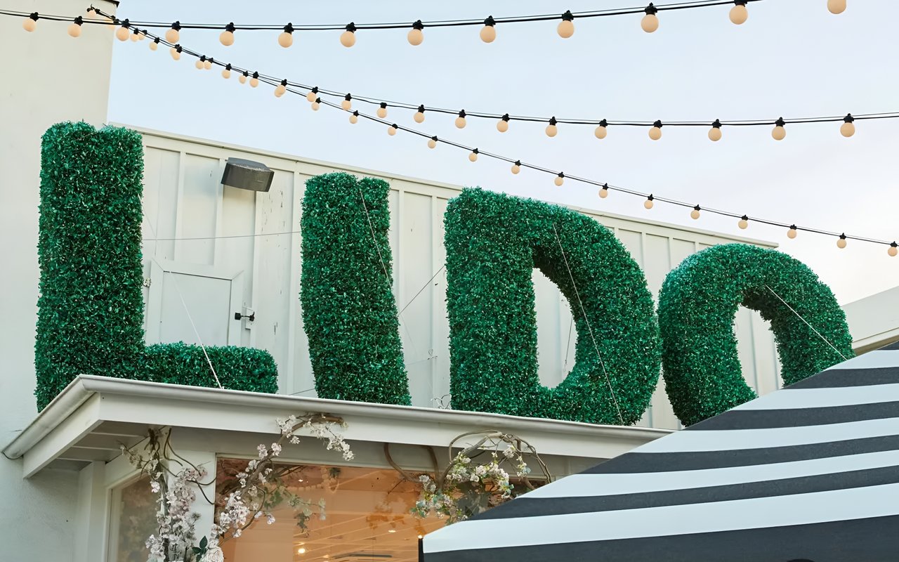  Large green letters, trimmed to look like hedges, spell out "LIDO" on the roof of a building with string lights overhead.