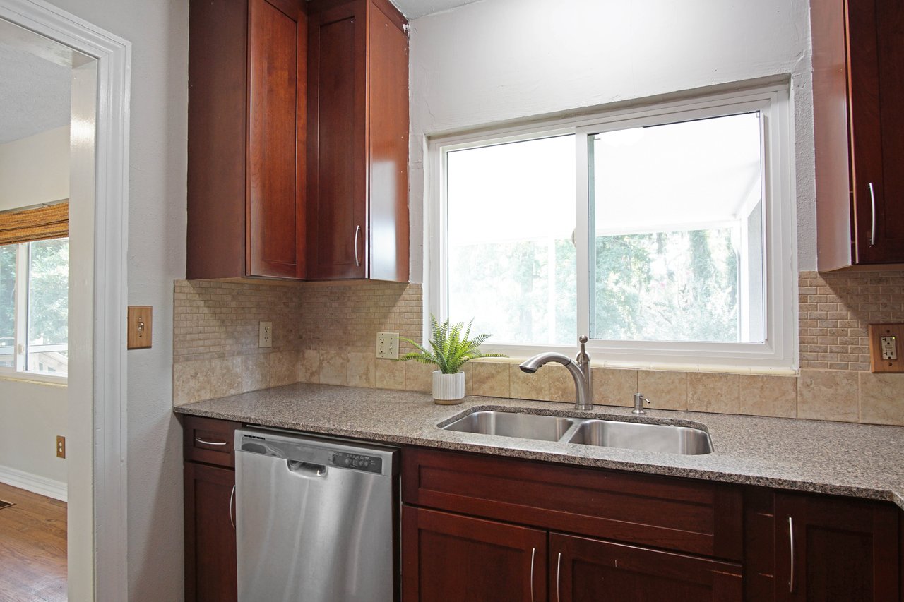 Bright kitchen with dark wood cabinets, granite countertop, and a double stainless steel sink. A potted plant sits by the window, adding a fresh touch.