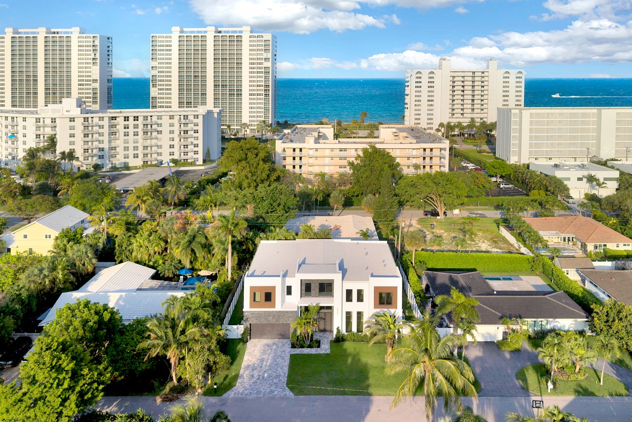 Modern two-story home amid palm trees, with oceanfront towers and the Atlantic Ocean behind it.