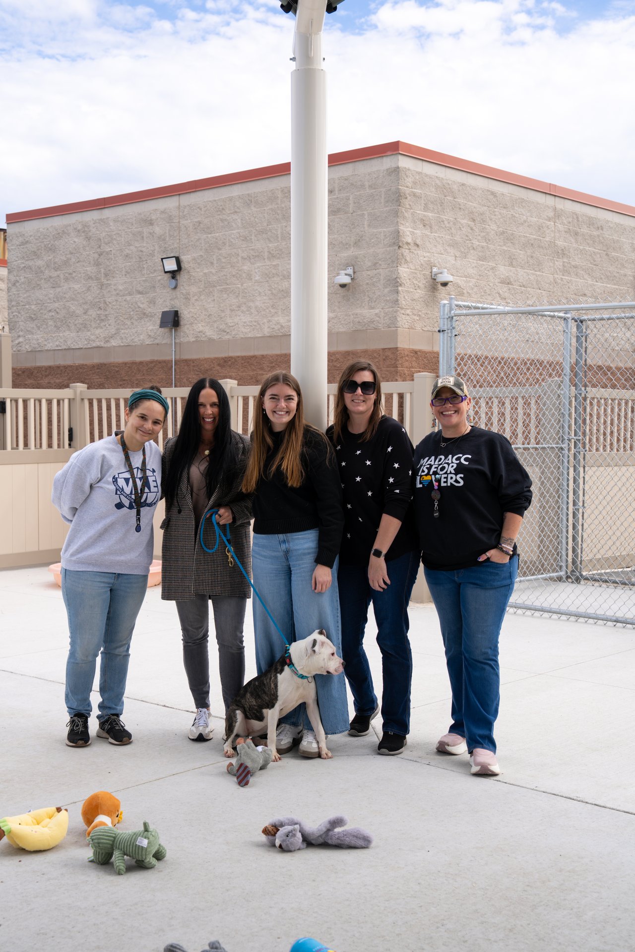 Five women pose with a leashed black-and-white dog among plush toys outside an animal shelter.