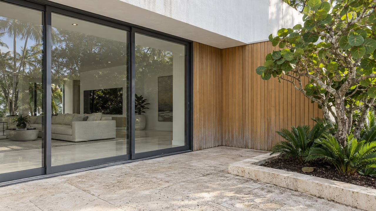 Contemporary home's limestone terrace beside tall closed glass sliders, warm wood cladding, and a sea-grape planting bed.