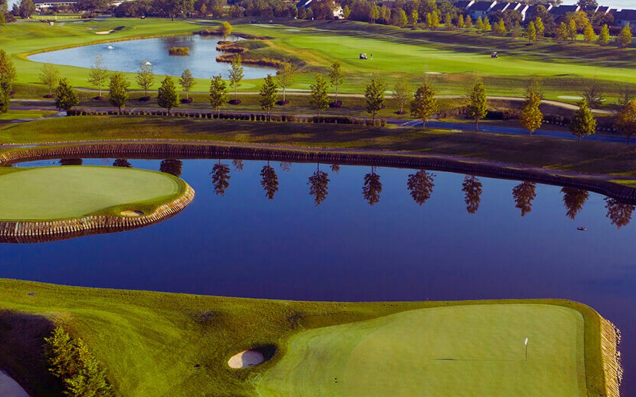 Golf course greens and fairways curve around a large pond reflecting autumn trees.
