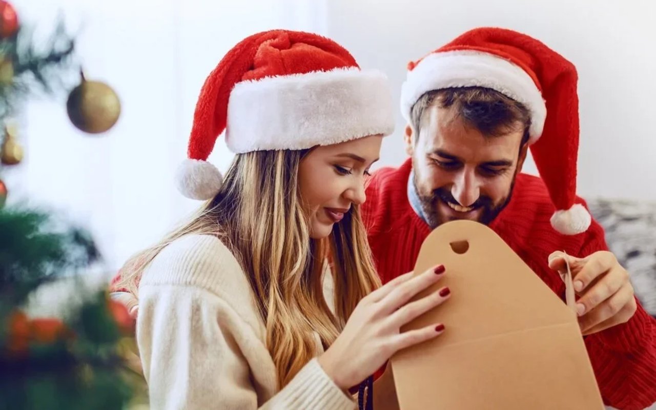 Couple in Santa hats open a cardboard box beside a decorated Christmas tree.