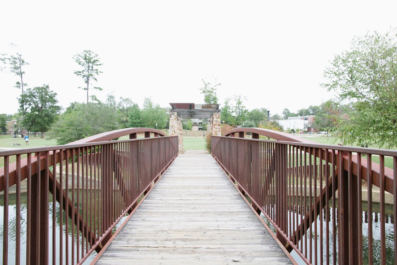 Wooden pedestrian bridge with red railings leading to a stone-and-wood arch in a park.