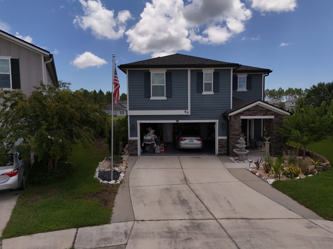 Two-story blue-gray house with an open garage, stone-accented porch, and American flag beside the driveway.