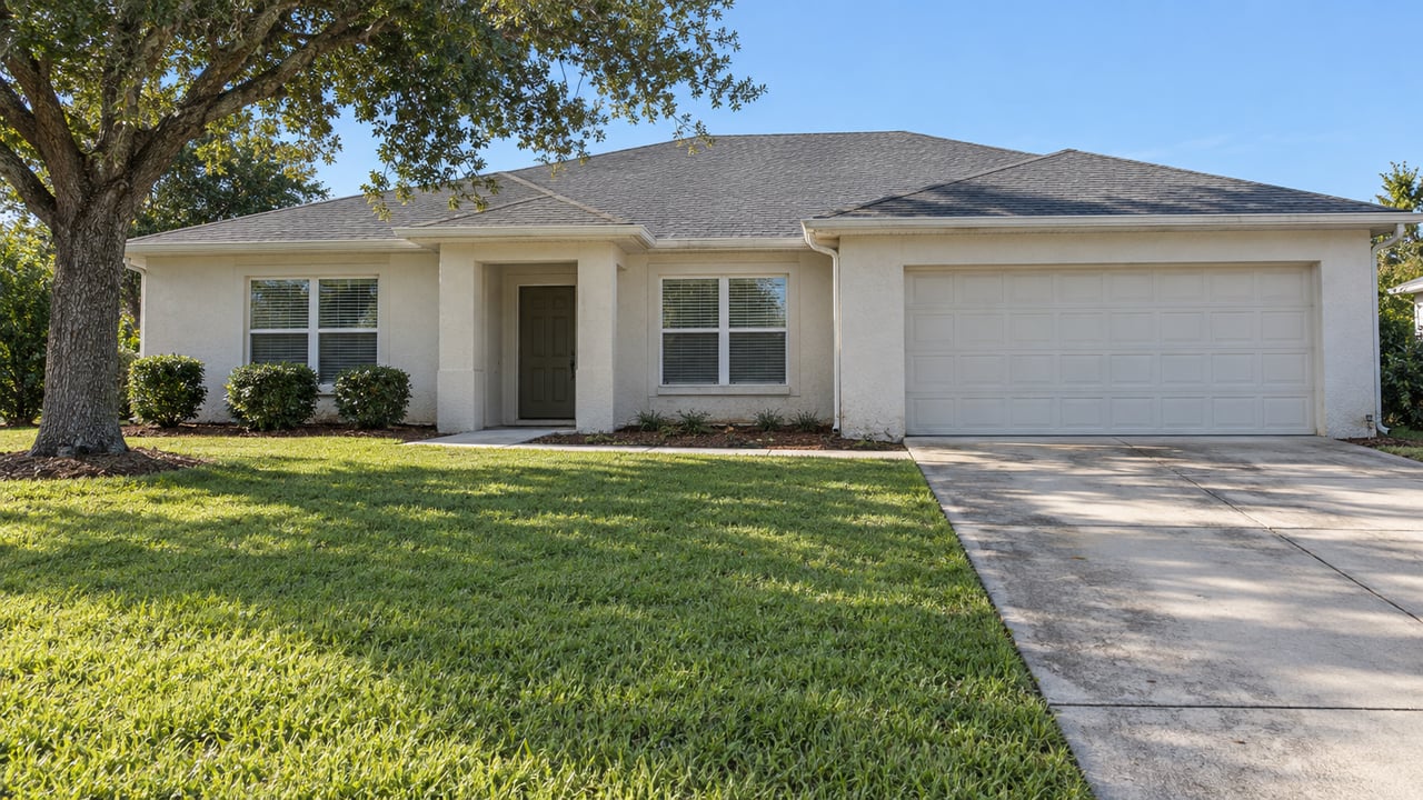 Ivory stucco single-story home with a charcoal hip roof, two-car garage, broad green front lawn and a spreading live oak.