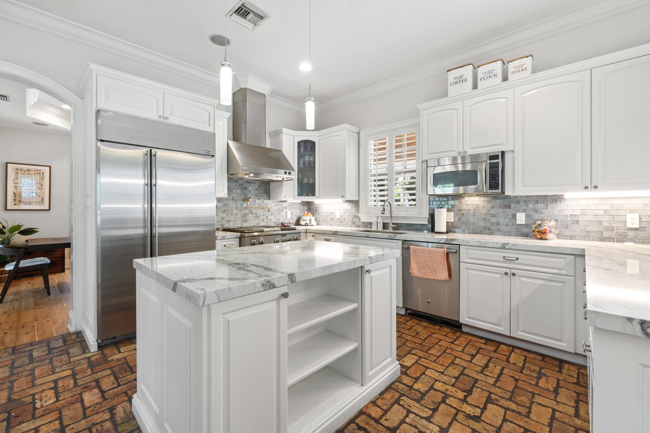 Bright white kitchen with a marble-topped island, stainless appliances, and brick flooring.