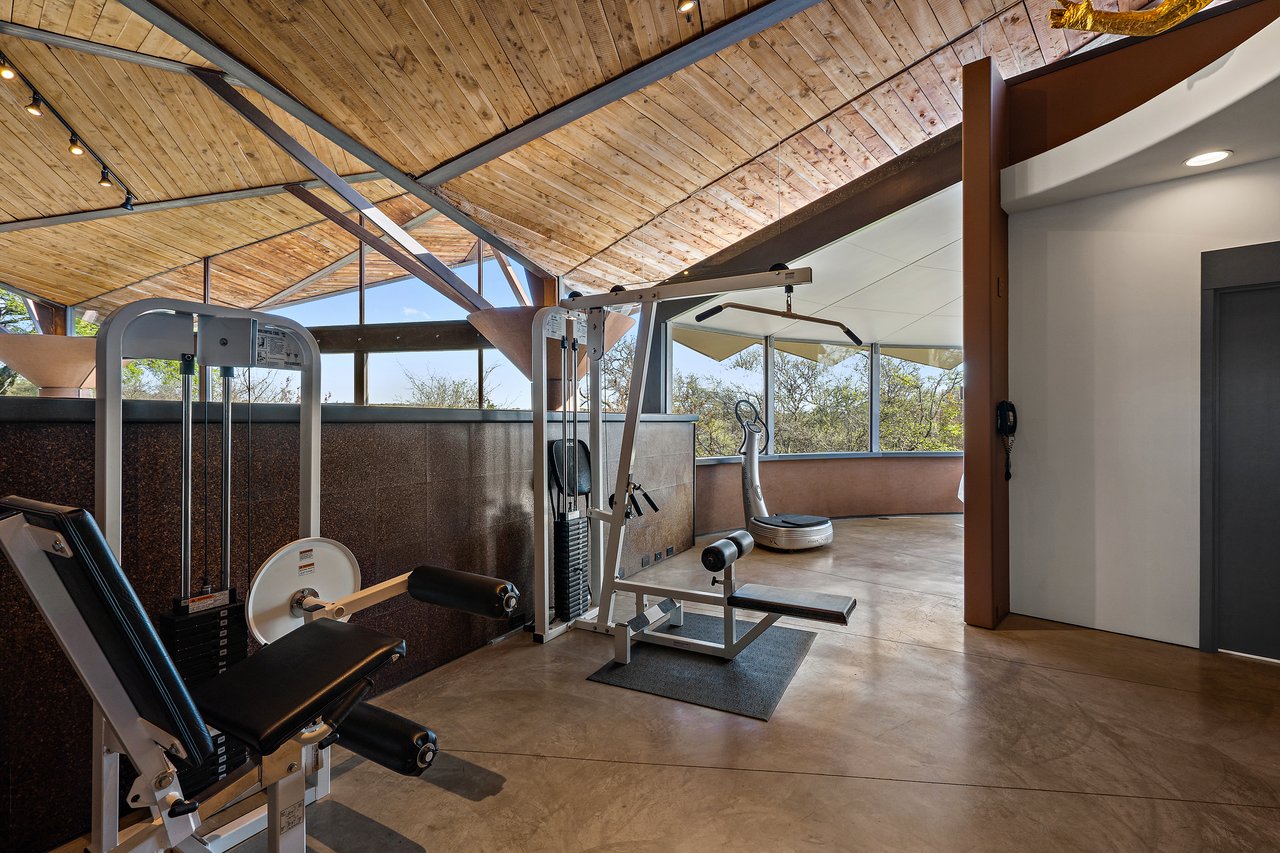 Home gym under a wood plank ceiling with exposed steel trusses and glass walls to the hill country, Lick Creek Lodge, Spicewood, Texas.