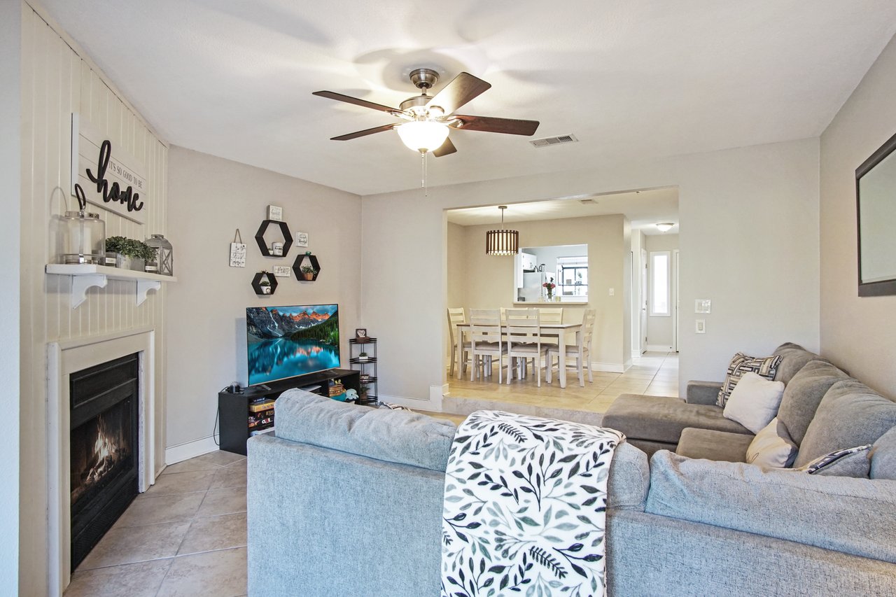 Cozy living room with a gray sectional sofa and patterned throw, facing a TV beside a fireplace. Wall decor and a dining area with wooden table are visible.