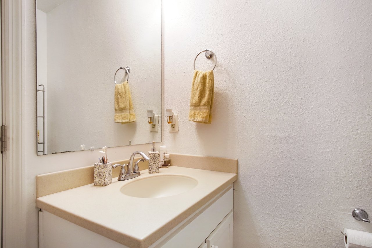 Small bathroom with white walls and beige countertop. A large mirror reflects a yellow hand towel hanging on a ring. The sink has a silver faucet.