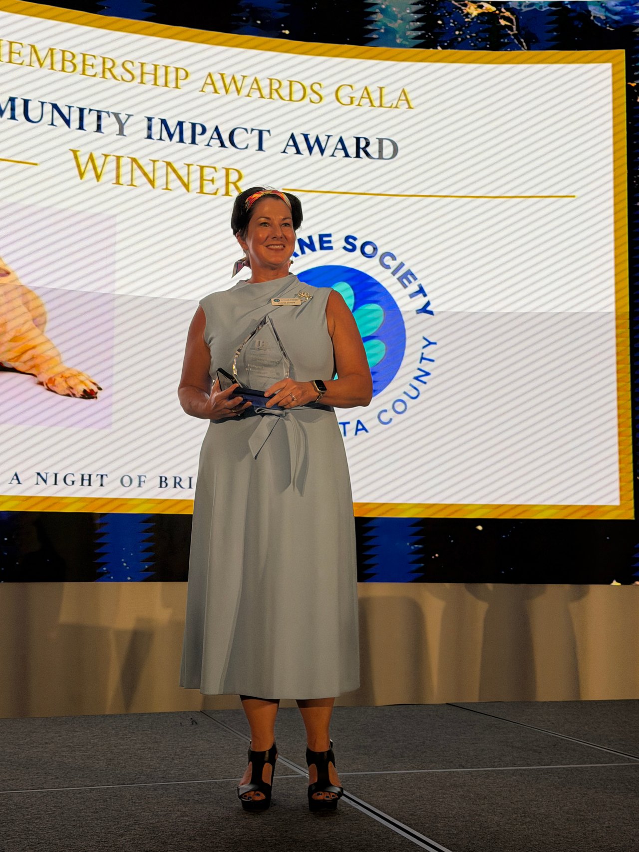 Woman in a gray dress holding a clear award in front of a Community Impact Award Winner backdrop.