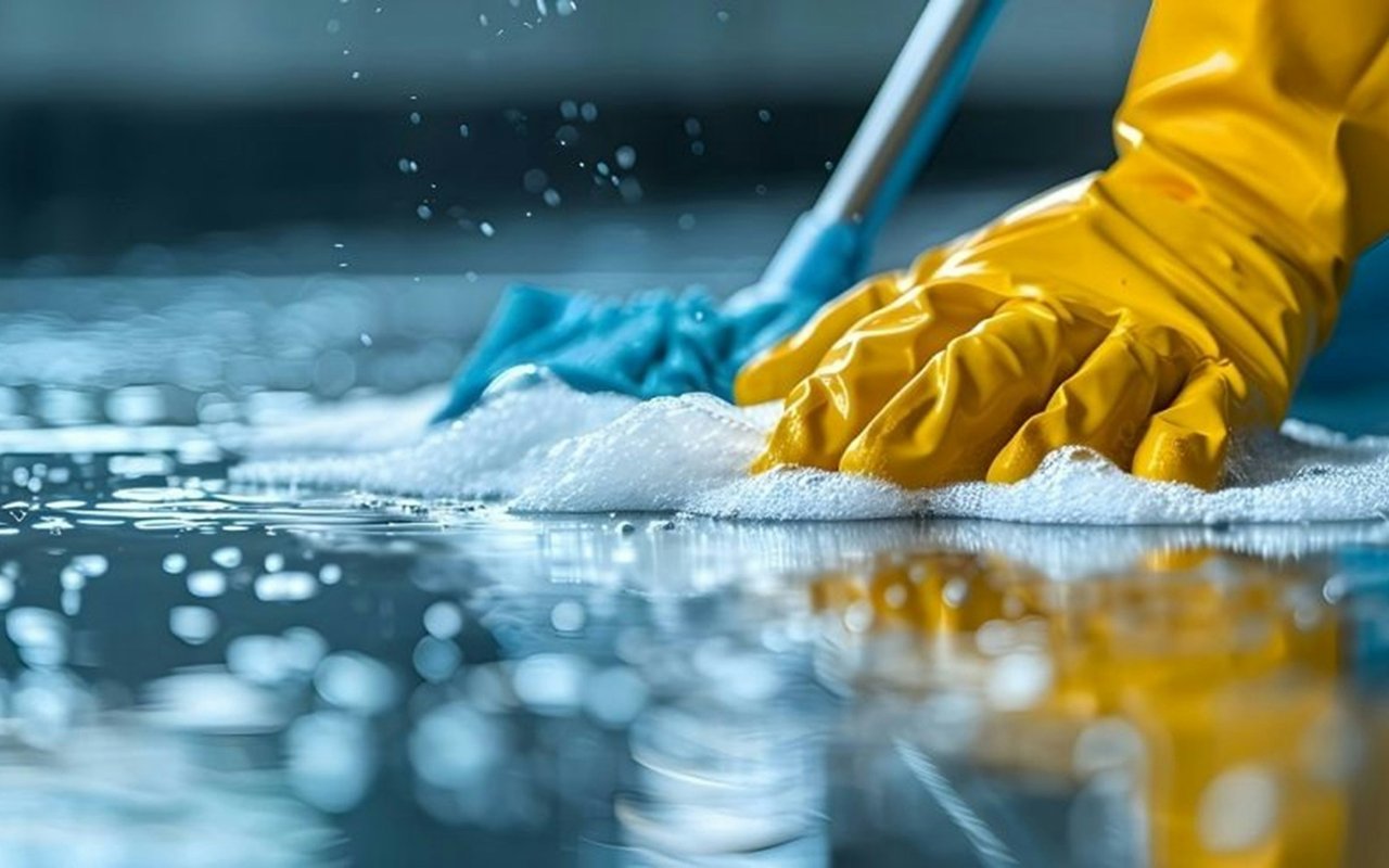 Yellow-gloved hand scrubbing a wet surface with a blue sponge amid soap suds.
