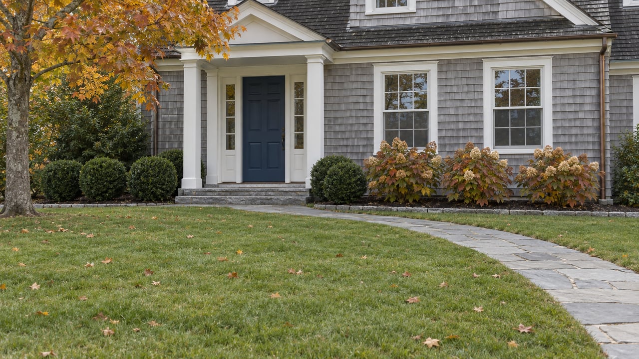 Cedar-shingled home with a blue front door, white portico, broad autumn lawn and amber maple leaves.