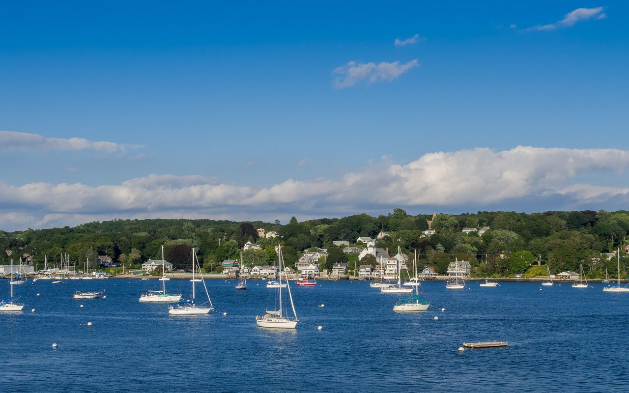Tiverton waterfront with sailboats in the harbor and homes along a wooded shoreline.
