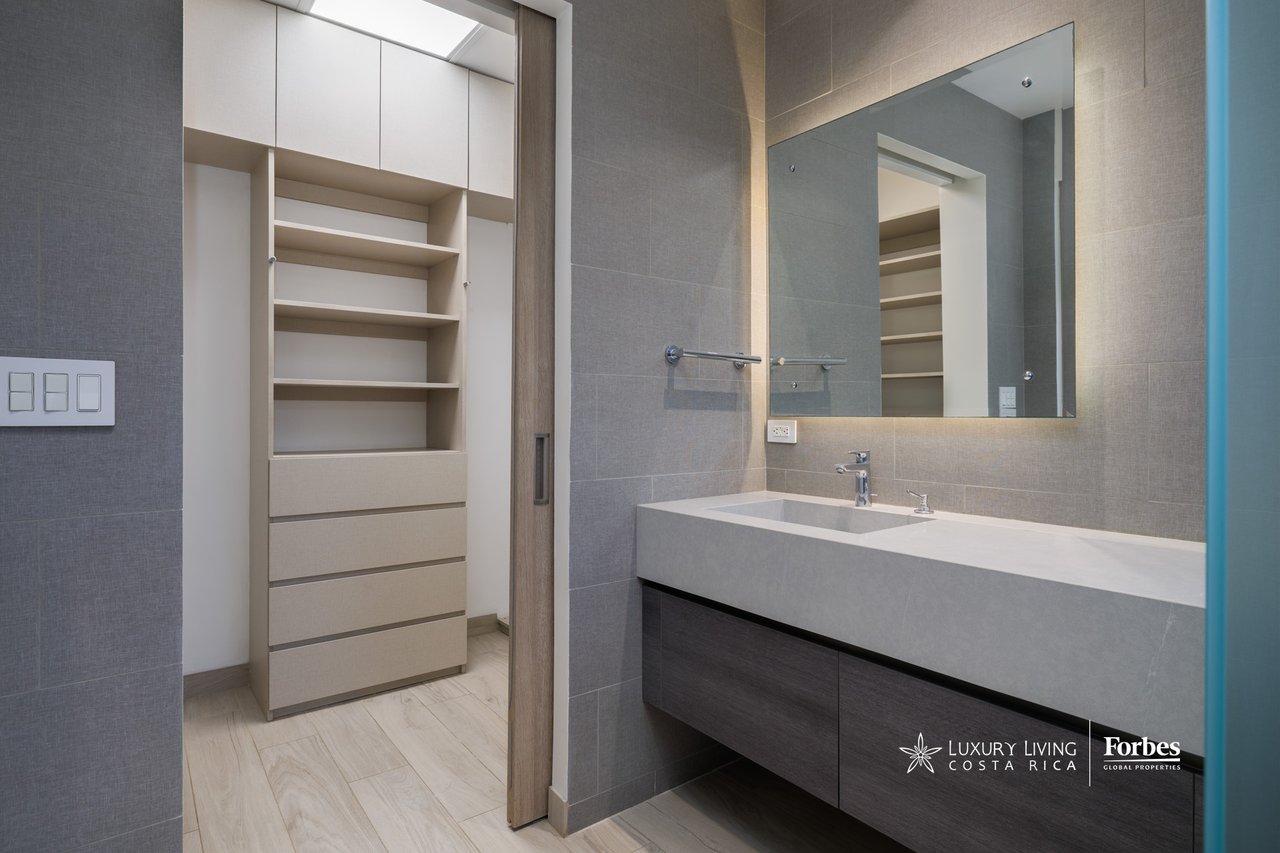 Bathroom vanity with illuminated mirror beside built-in open shelving and drawers.
