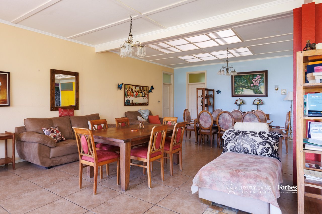 Living and dining room with wooden furniture, red chair cushions, tiled floors, and colorful wall art.