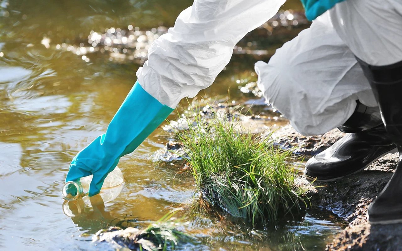 Person in protective clothing retrieves a plastic bottle from a shallow, sunlit stream.