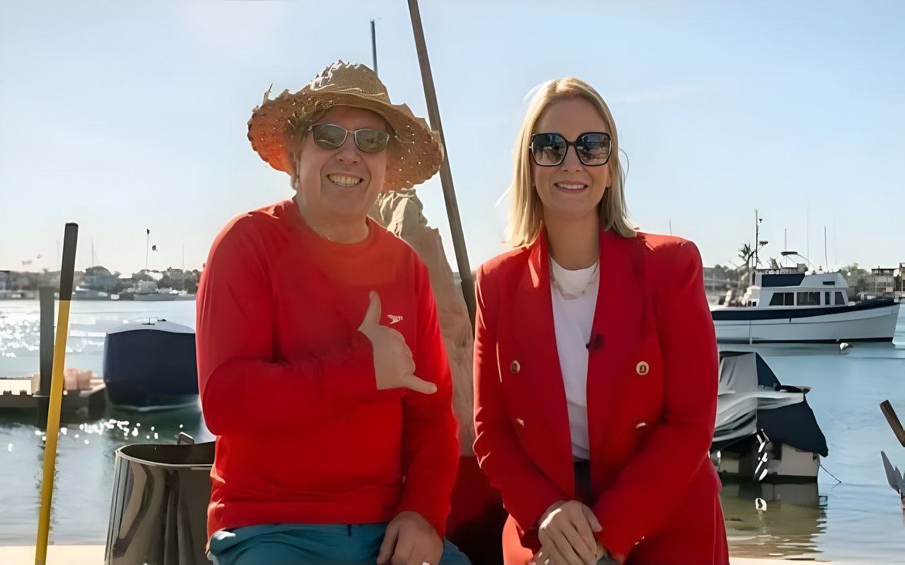 A man in a straw hat and a woman in sunglasses, both wearing red jackets, sit by the water at a marina.