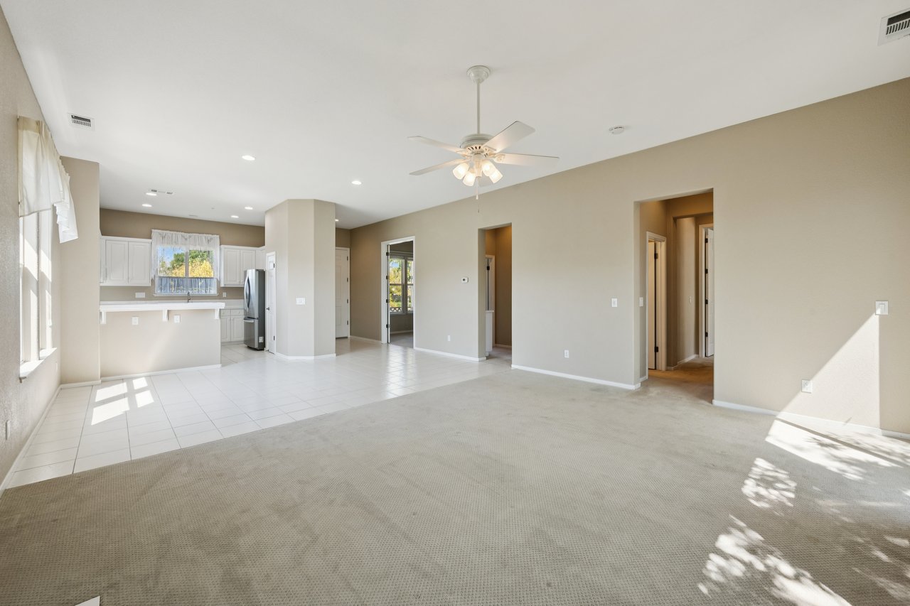 Vacant living room opening to a white-cabinet kitchen, with beige carpet and a ceiling fan.