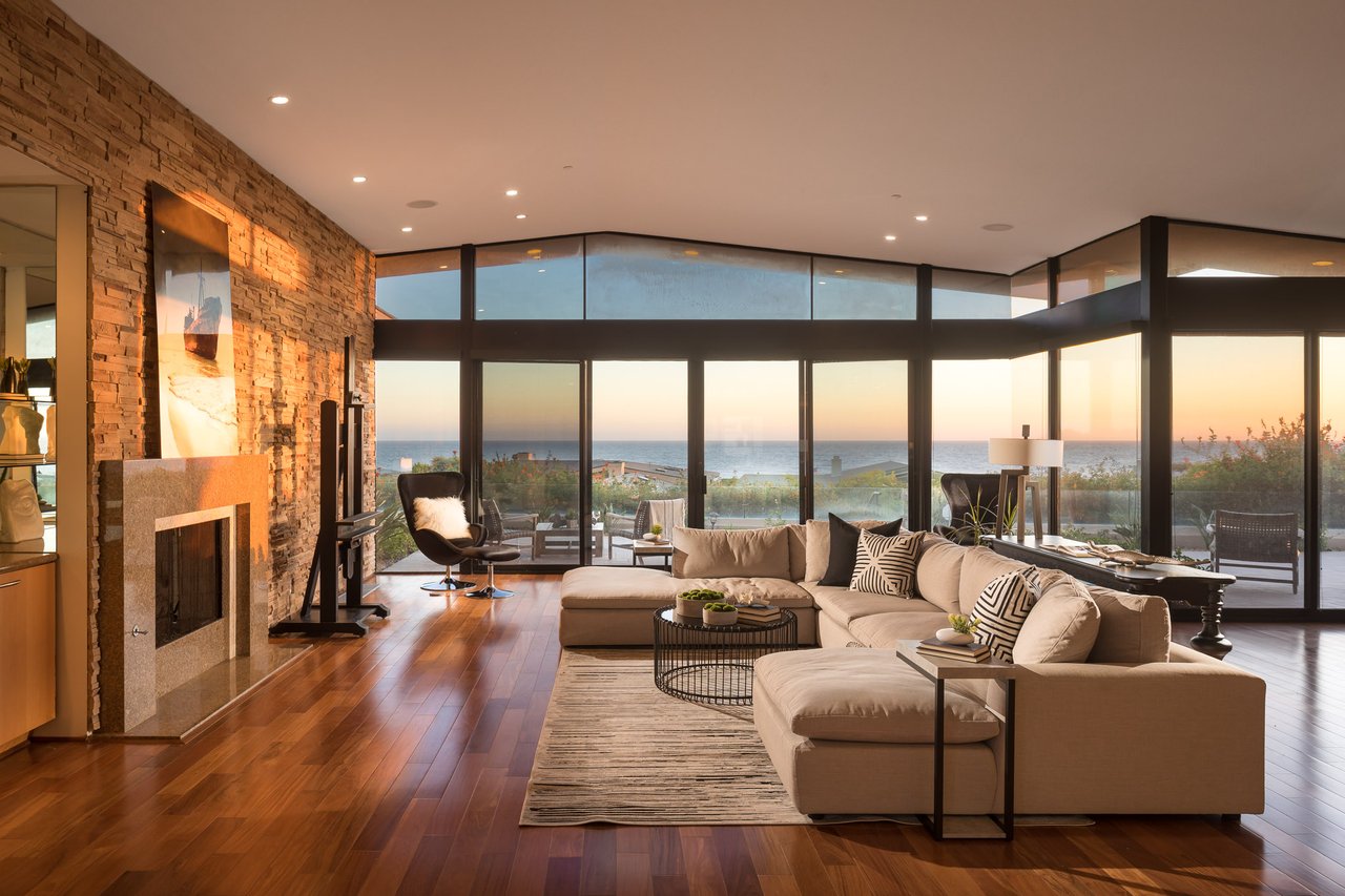 Contemporary living room with a stone fireplace, sectional sofa, and sunset ocean views through tall windows.