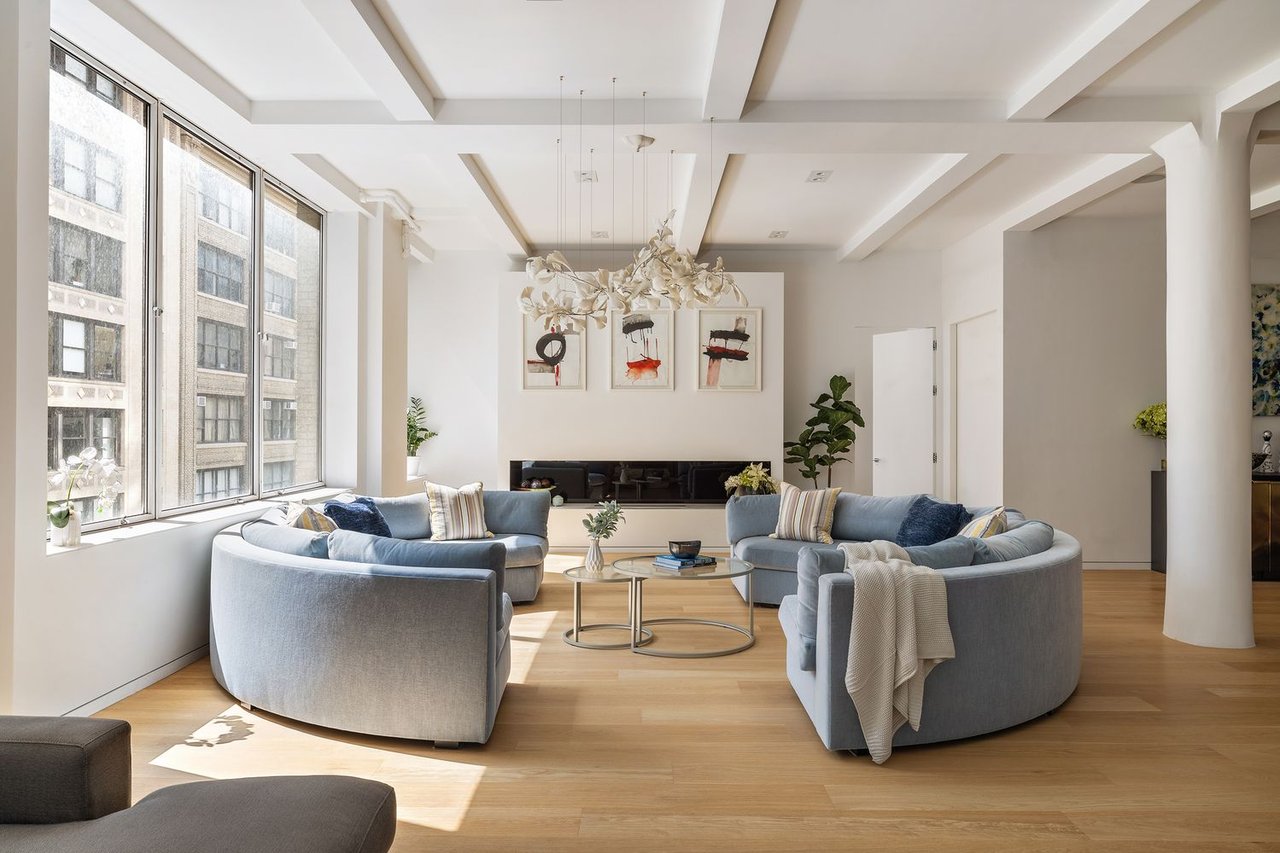 Bright living room with curved blue-gray sofas, tall city-facing windows, and a sculptural chandelier.