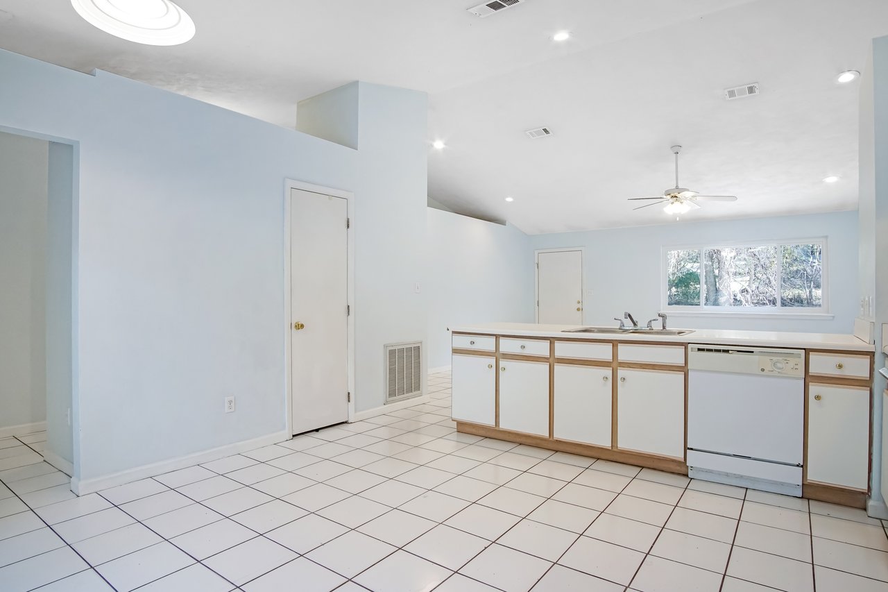 A spacious, empty kitchen with light blue walls, white tiled floor, and a central island. A ceiling fan and large window evoke a bright, airy feel.