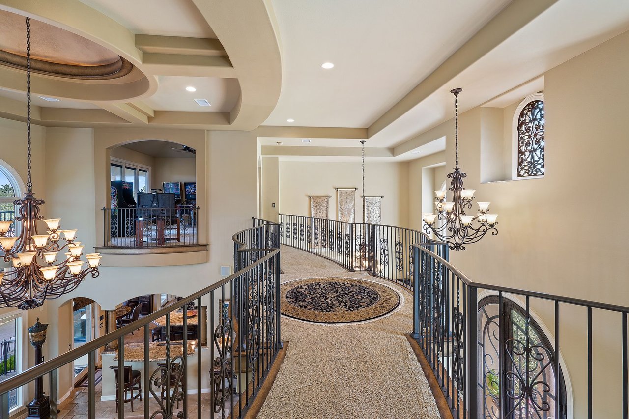 Upstairs gallery hallway with wrought iron railing, a cove ceiling, and chandeliers at 25715 Cliff Circle, Lakecliff on Lake Travis, Spicewood, Texas.