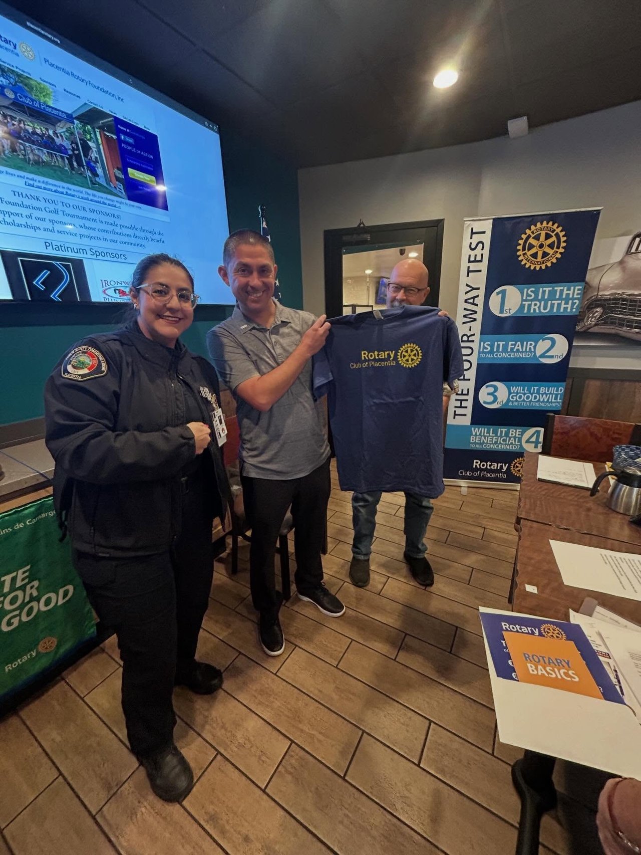 Three adults pose with a Rotary Club of Placentia shirt beside Rotary banners.