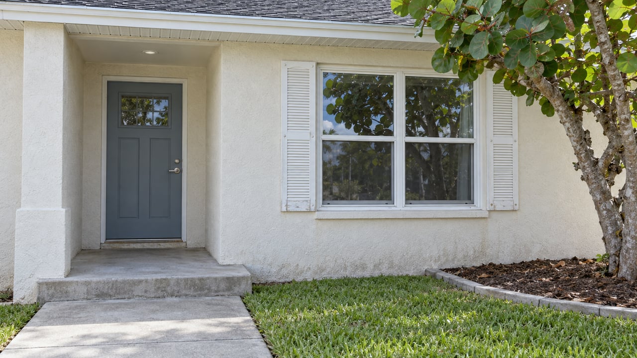 Close view of a cream stucco home's recessed blue-gray door, white-trimmed window and weathered concrete porch beside a sea-grape tree.