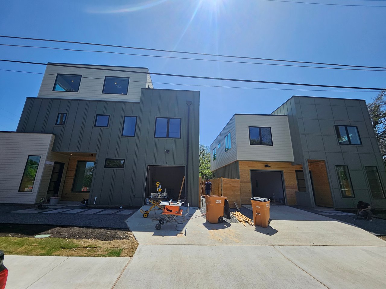 Modern multi-story homes under construction, with dark green siding and building materials in the driveway.
