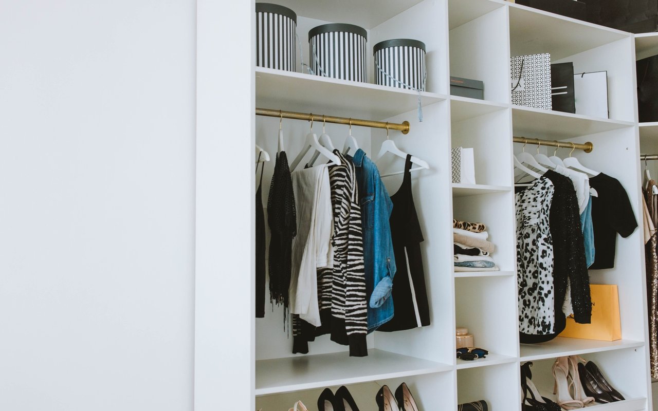 Walk-in closet with white built-in shelving, brass clothing rods, organized garments, shoes, and storage baskets.