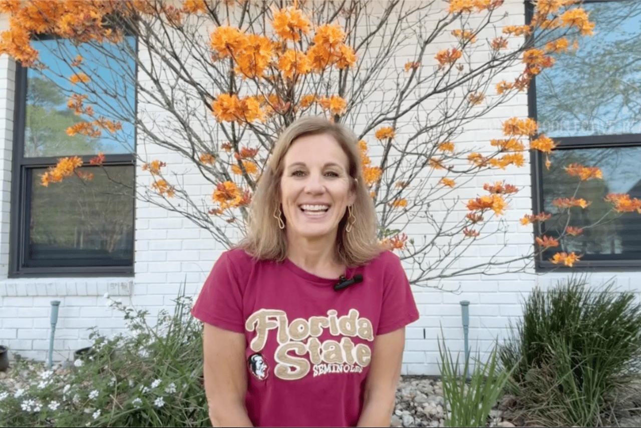 Smiling woman in a Florida State Seminoles shirt stands before a white brick house and orange-leaved tree.