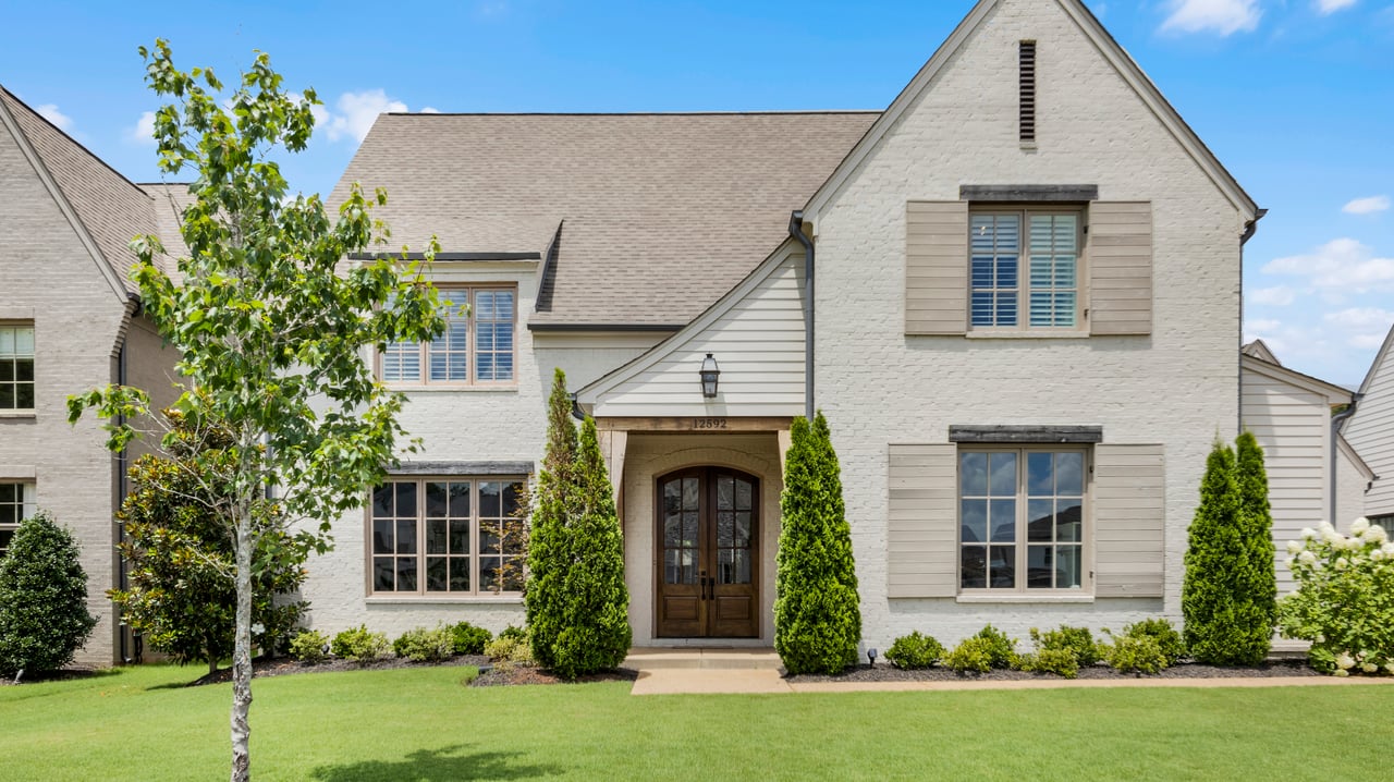 White brick two-story home with taupe shutters, dark double doors, and a landscaped front lawn.