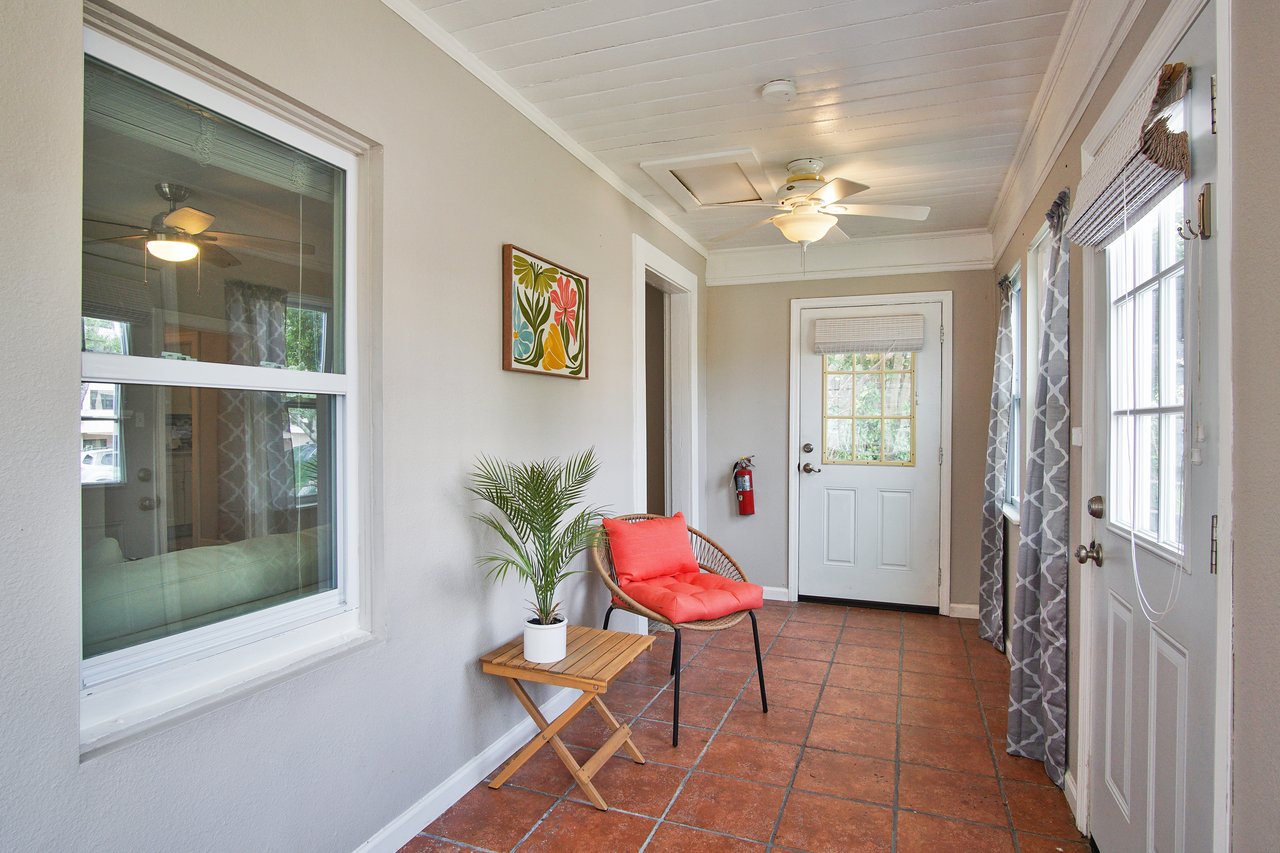 A bright sunroom with terracotta tiles, a wicker chair with a red cushion, and a small plant on a table. A vibrant painting hangs on a gray wall.