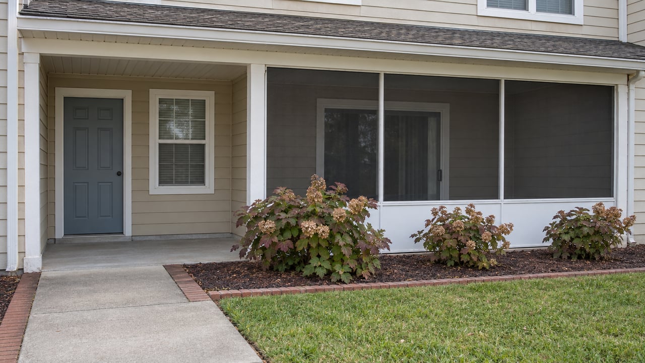 Sand-colored condominium exterior with a blue-gray entry door, screened porch, white trim, and oakleaf hydrangeas beside a concrete approach.