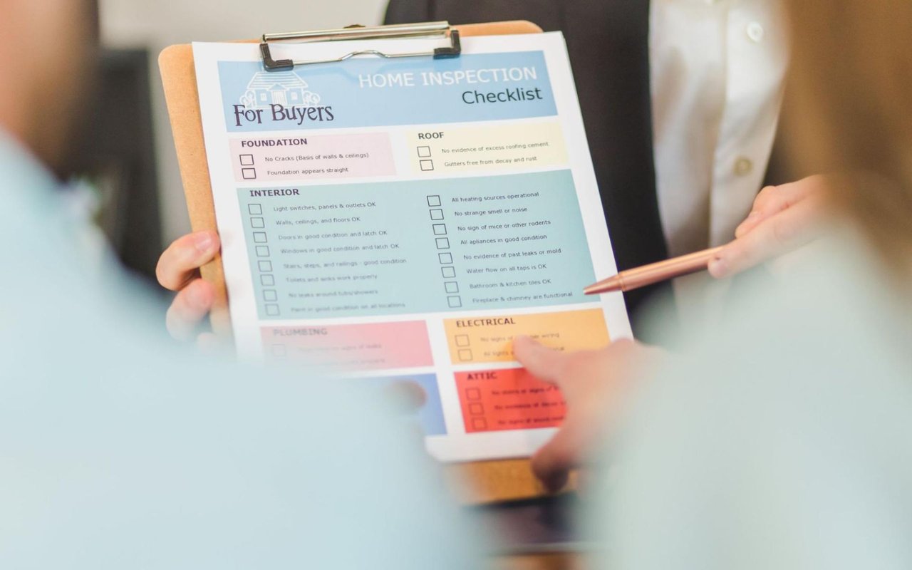  A close-up view shows a person's hands holding a clipboard with a "HOME INSPECTION Checklist For Buyers" form. 