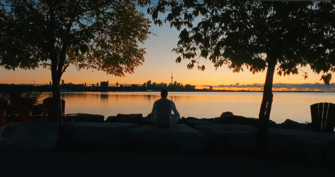 Person seated beneath trees by the water, watching the Toronto skyline at sunset.