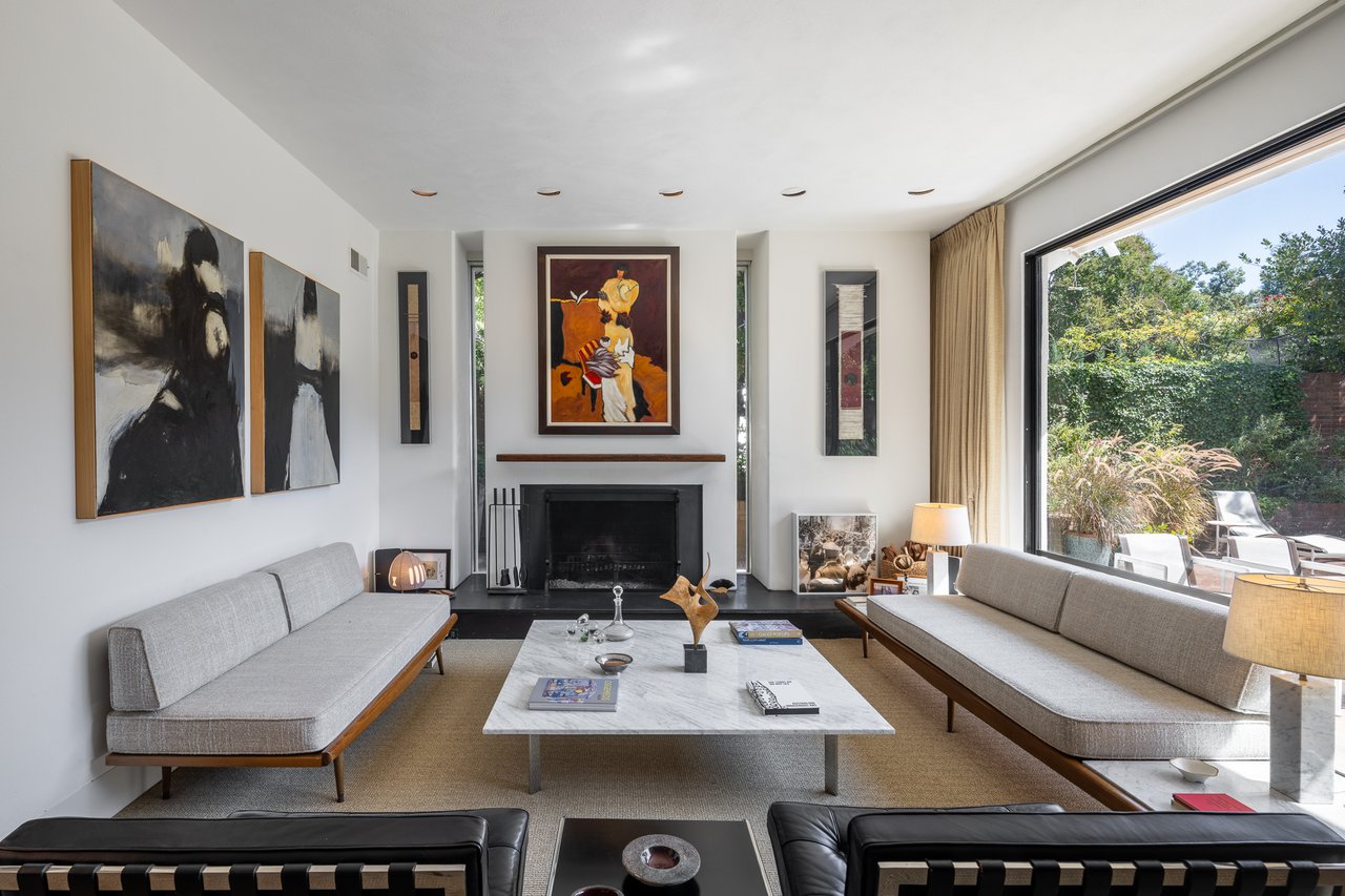 Modern living room with gray sofas, a fireplace, abstract art, and a large window overlooking a garden.