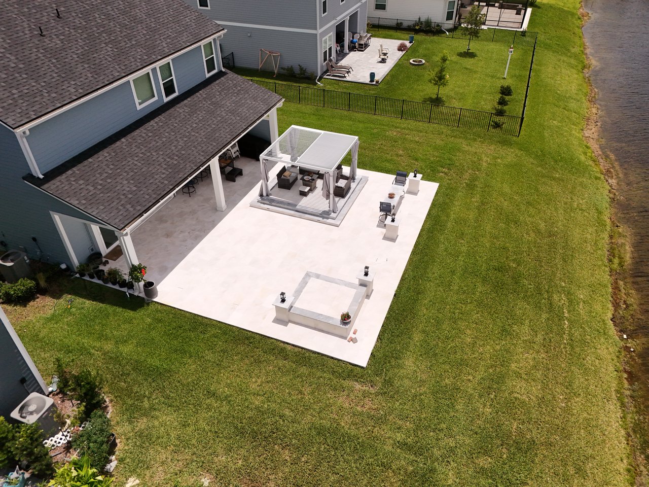 Aerial view of a gray home’s spacious patio with a white pergola beside a waterway.