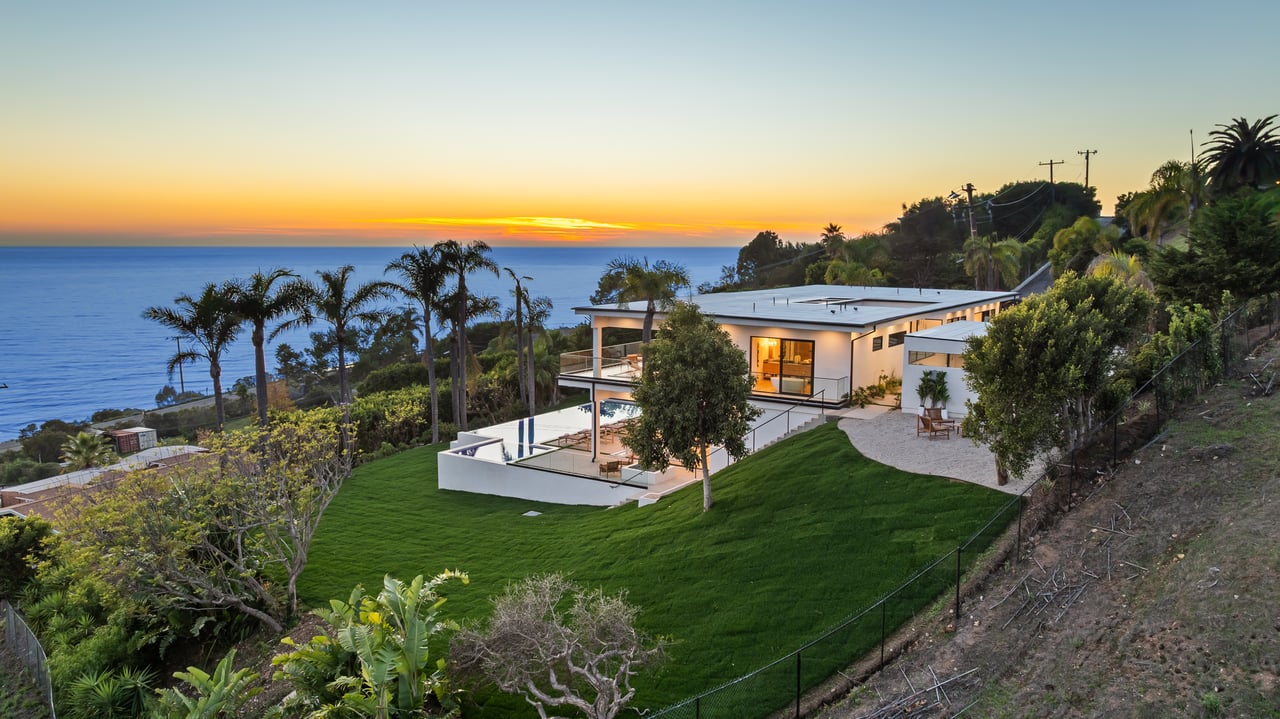Modern coastal home overlooking the ocean at sunset, with a green lawn, palm trees, and a small pool.