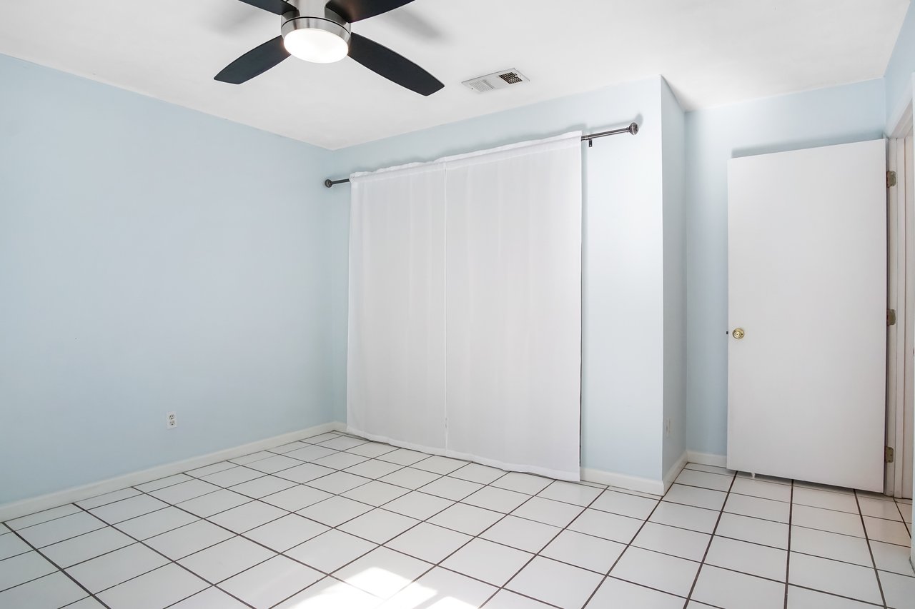 Empty room with pale blue walls and white tiled floor. A white curtain covers a window, and a white door is slightly open. A ceiling fan is on top.