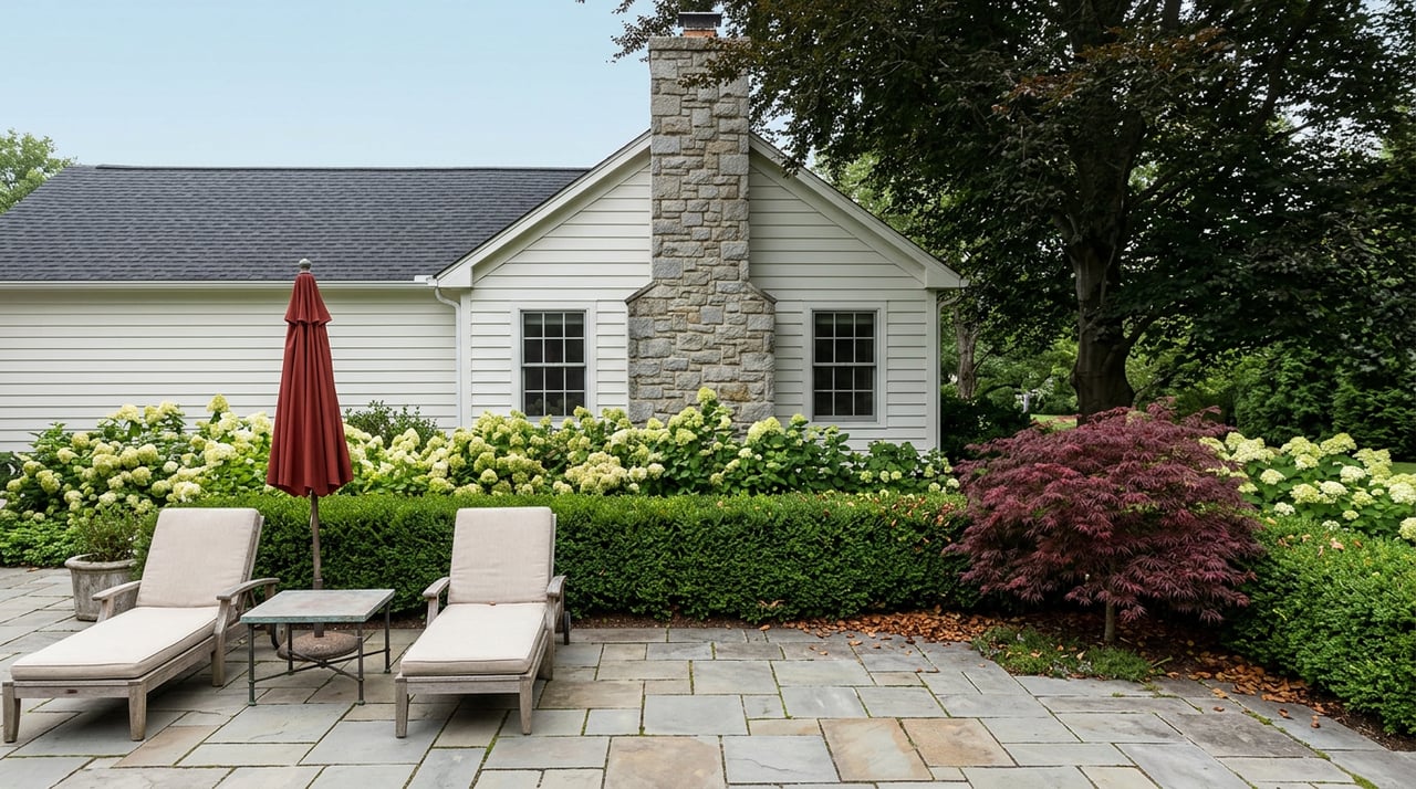 Bluestone terrace with teak lounge chairs and a red umbrella, bordered by boxwood and hydrangeas beneath a beech tree.