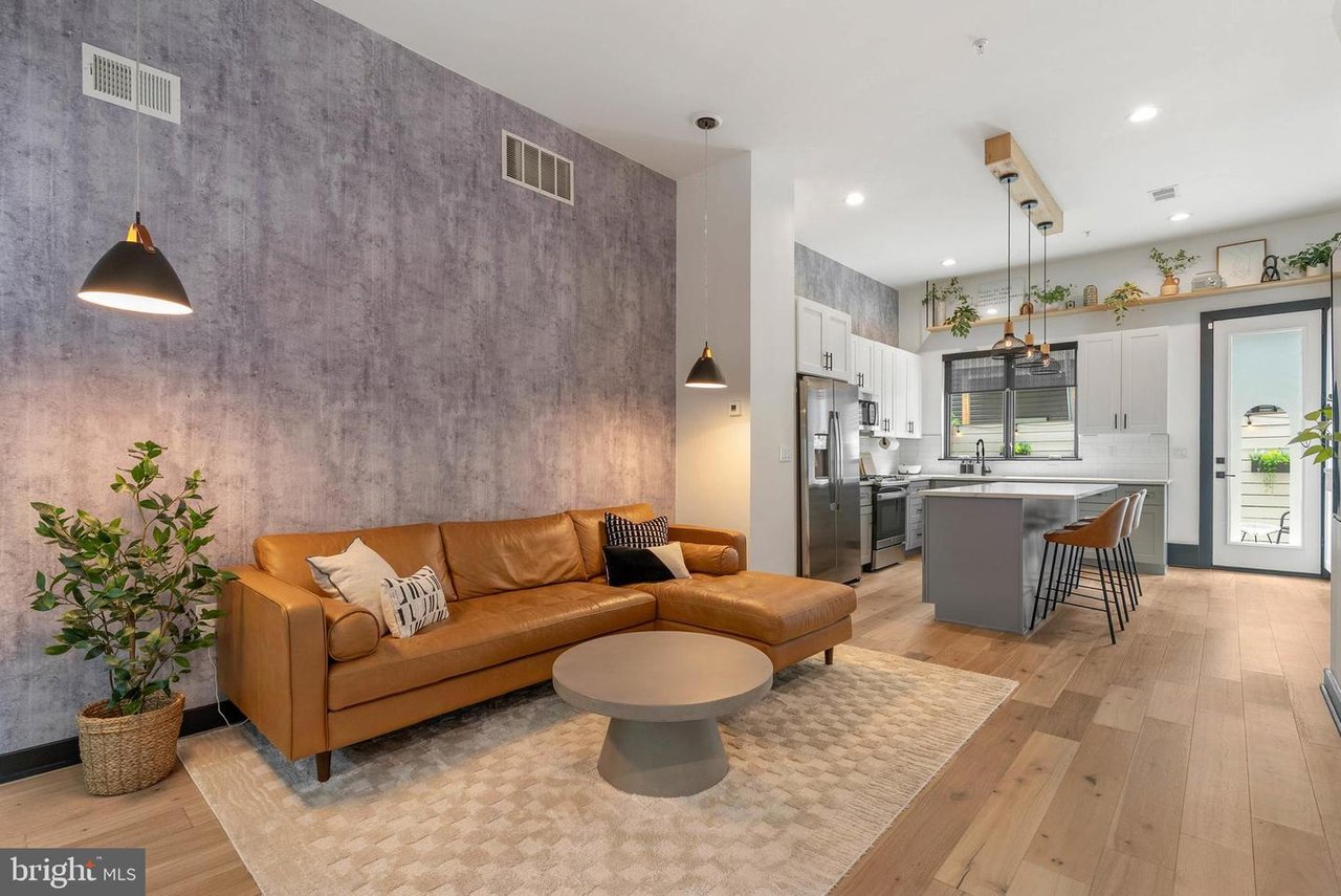 Open-plan living room and kitchen with a caramel leather sectional and textured gray accent wall.