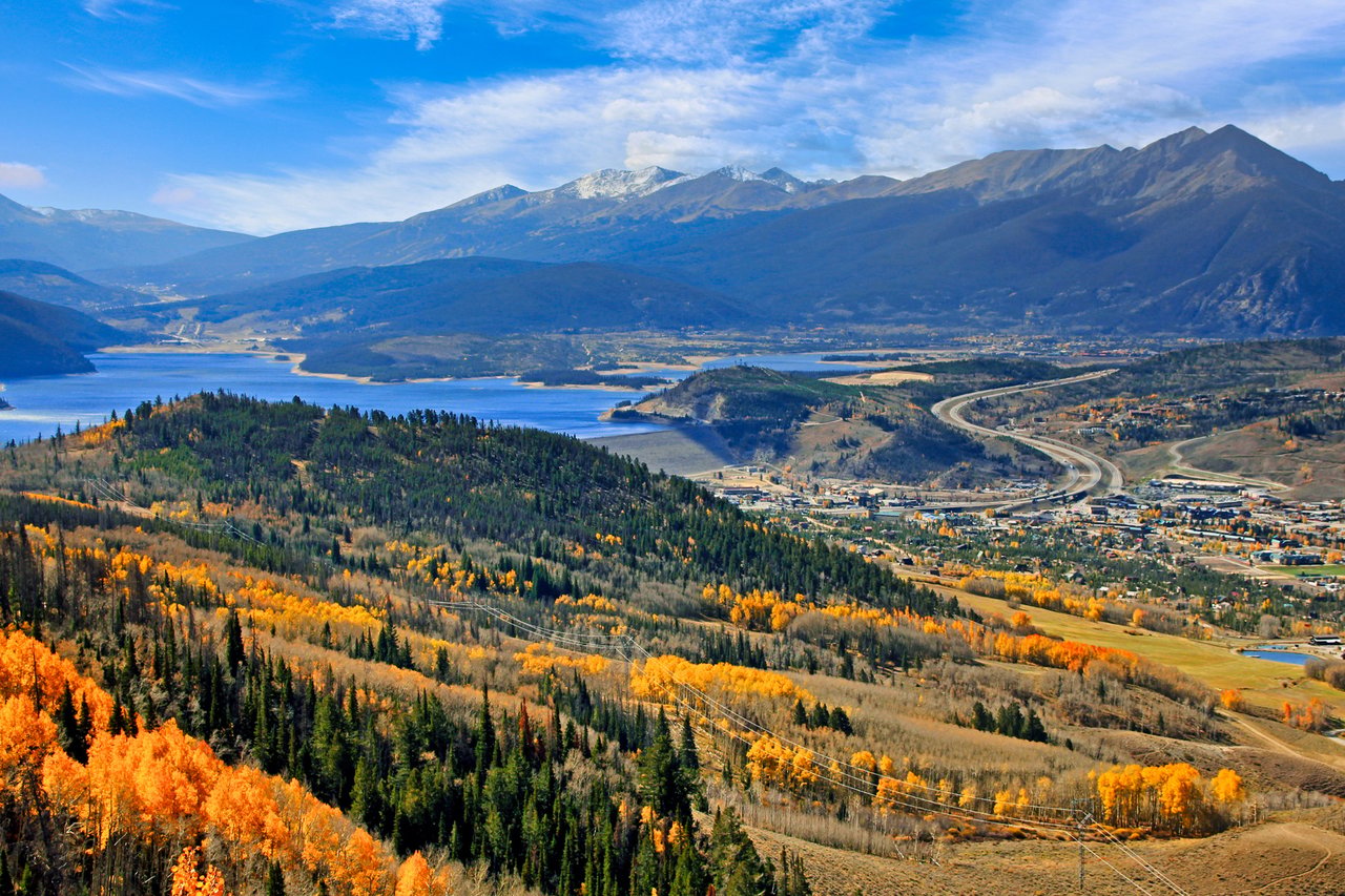Autumn mountain valley with a reservoir, forested slopes, and a winding highway beneath high peaks.