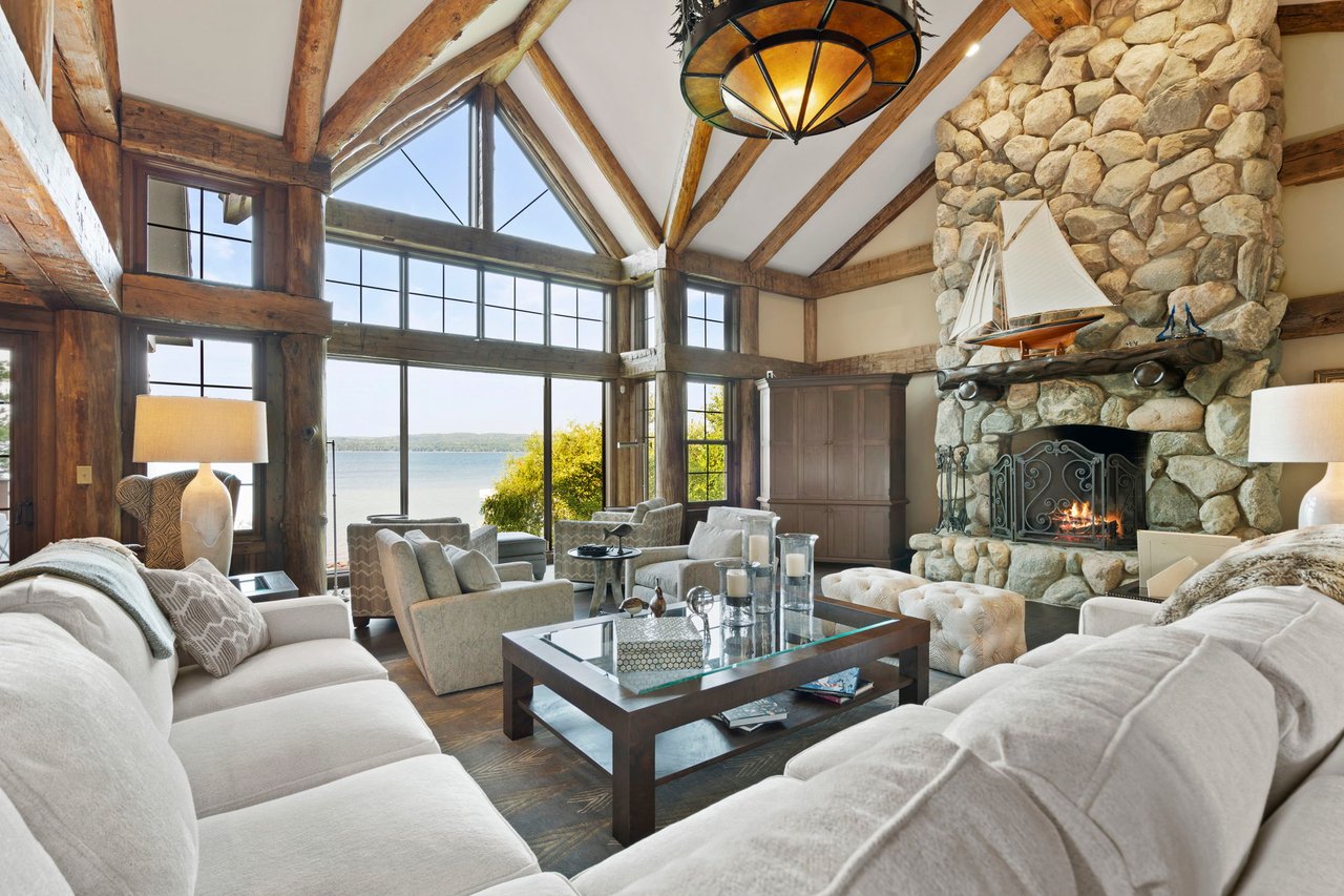 Living room with exposed timber beams, a stone fireplace, and large windows overlooking the water.